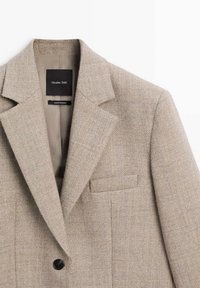 Blazer in misto lana color tan con revers a punta, chiusura a un bottone e taschino sul petto. Presenta un tessuto testurizzato con un sottile motivo a spina di pesce.