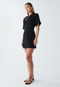 Črna kratka obleka s ovratnikom, kratkimi rokavi in dvema sprednjima žepoma. Tkanina se zdi gladka, zasnova pa je prilegajoča.