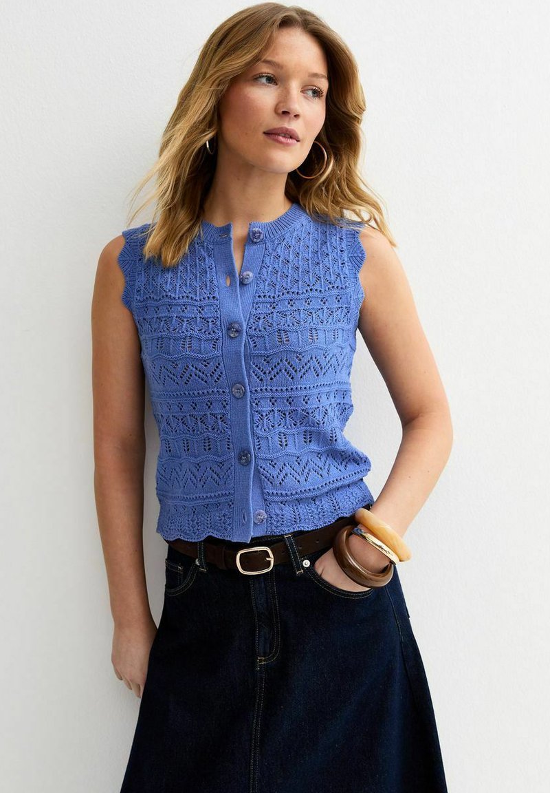 Femme portant un haut tricoté bleu sans manches avec un motif en dentelle, une jupe foncée et de grands bracelets, debout contre un mur blanc.