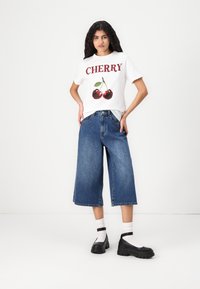 Balta T-kreklu ar sarkano "CHERRY" tekstu un ķiršu grafiku. Zili platajiem denima bikšiem. Melni platformas apavi ar potītes siksniņu.