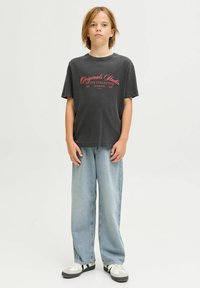 Niño de pie, vistiendo una camiseta gráfica negra con texto rojo, jeans ancho de tela vaquera azul claro y zapatillas blancas con detalles en negro.