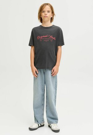 Niño de pie, vistiendo una camiseta gráfica negra con texto rojo, jeans ancho de tela vaquera azul claro y zapatillas blancas con detalles en negro.
