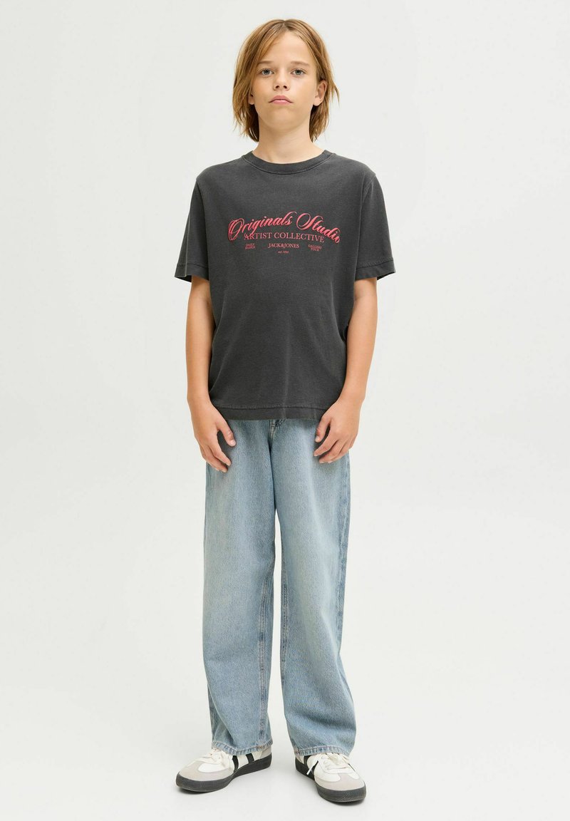 Niño de pie, vistiendo una camiseta gráfica negra con texto rojo, jeans ancho de tela vaquera azul claro y zapatillas blancas con detalles en negro.