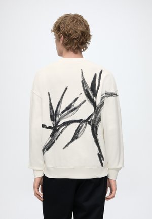 Personne blonde portant un sweat-shirt blanc avec un motif végétal abstrait en coups de pinceau noirs dans le dos, associé à un pantalon noir.