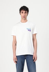 Hvid bomulds T-shirt med korte ærmer, prydet med et flerfarvet grafisk design på venstre brystområde. Båret med blå denim jeans.