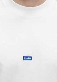 Valge ümara kaelusega särgi lähivõte, millel on väikene sinine ristkülikukujuline silt „HUGO“ rinnal.