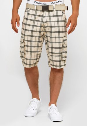 Pantalones cortos de carga de cuadros beige con líneas negras, bolsillos laterales y un cinturón beige. Llevados con zapatillas blancas, mostrando un ajuste relajado.
