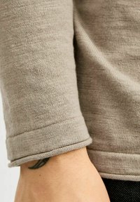 Manica di un maglione lavorato a maglia beige con polsino arrotondato. Il tessuto è morbido con una leggera texture. Un tatuaggio astratto è visibile sul polso.