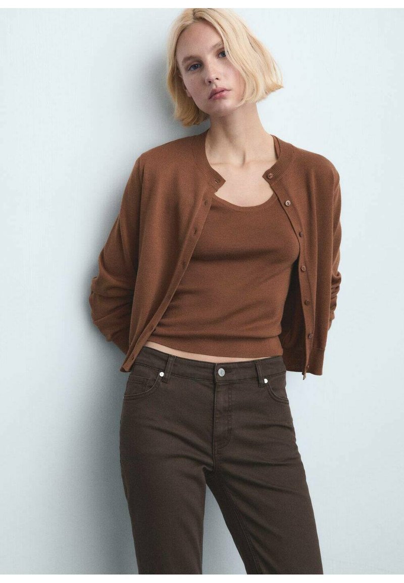 Jeune femme aux cheveux blonds courts portant un cardigan marron boutonné sur un débardeur marron et un pantalon marron foncé, se tenant contre un mur blanc.