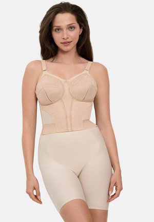 Femme aux longs cheveux bruns portant un bustier en dentelle beige et un short gainant taille haute, debout devant un fond clair uni.