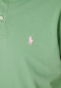 Zelené polo tričko vyrobené z texturované látky, s knoflíkovou légou na tři knoflíky a růžovou vyšívanou nášivkou koně a jezdce na hrudi.