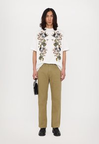Hvid oversized T-shirt med blomstermønster, parret med olivengrønne strukturerede bukser. Tilbehøret inkluderer en lille sort taske. Sorte sko.
