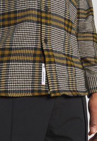 Chemise à carreaux boutonnée en noir, blanc et jaune moutarde avec une étiquette de marque blanche visible, portée avec un pantalon noir et une vue partielle d'une main.