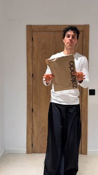Giovane uomo con camicia bianca e pantaloni neri che lancia un pacco marrone di Zalando davanti a una porta di legno in una stanza luminosa.