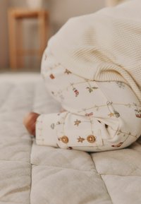 Balta kūdikių drabužių medžiaga su šventiniu raštu, kuriame pavaizduoti ornamentai, žvaigždės ir saldžiųjų lazdelių. Yra mediniai mygtukai ant rankogalių, drabužiai padėti ant tekstūruoto prijuostės.