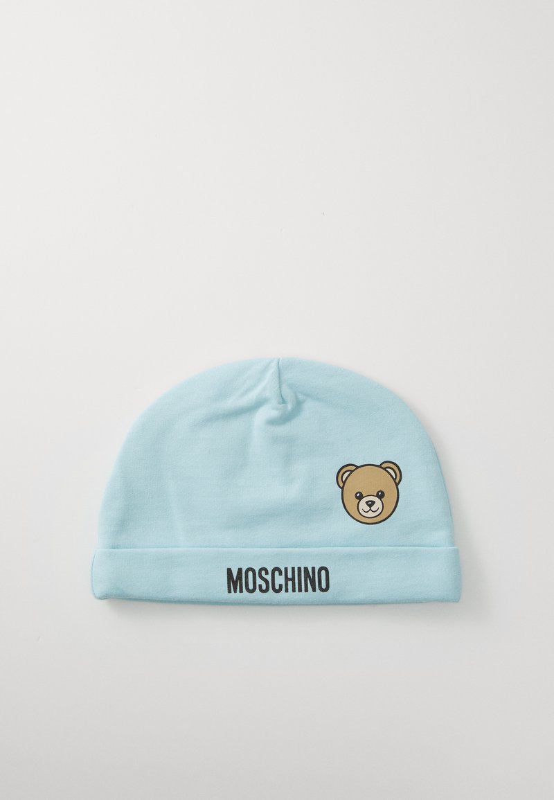 Šviesiai mėlynas kūdikių dangtelis su meškiuko veido grafika ir užrašu "MOSCHINO" ant sulankstymo krašto, padėtas ant baltos fono.