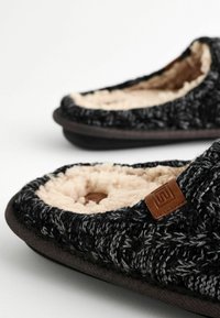 Knitted black and gray slippers with a soft beige lining, featuring a durable dark sole and a small brown leather accent.
