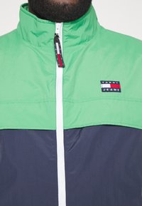 Farbenblockierte Jacke in leuchtendem Grün und Marineblau, hergestellt aus leichtem Material. Verfügt über einen hohen Kragen und einen zentralen Reißverschluss. Logopatch auf der Brust.