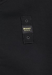 Svart tyg med en sl ät textur har en svart etikett som är sydd på, där det står "Blauer!" och "U.S.A." i gult, med silveröljetter.