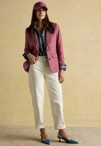 Žena v ružovom saku a čiapke, námornícka pruhovaná košeľa, slonovinovo biele rifle s vyhrnutými manžetami a tyrkysové sandále na podpätku, stojí pri béžovej stene.