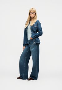 Femme blonde portant des lunettes, une chemise bleu clair, une veste en denim bleu foncé, un jean à jambes larges et des chaussures pointues marron, debout devant un fond blanc.