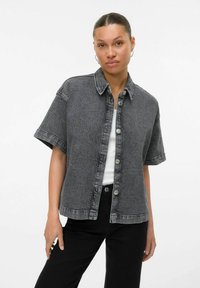 Camicia in denim grigio scuro con maniche corte, chiusura con bottoni, colletto e dettagli di cucitura sottili. Abbinata a una maglietta bianca e jeans neri.