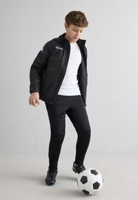 Jongen in zwarte sportkleding en voetbalschoenen die een zwart-witte voetbal onder controle houdt op een lichte vloer tegen een witte muur.