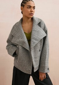 Gray wool-blend oversized jacket with a notched collar, deep pockets, and button accents, layered over a green ribbed tank top.