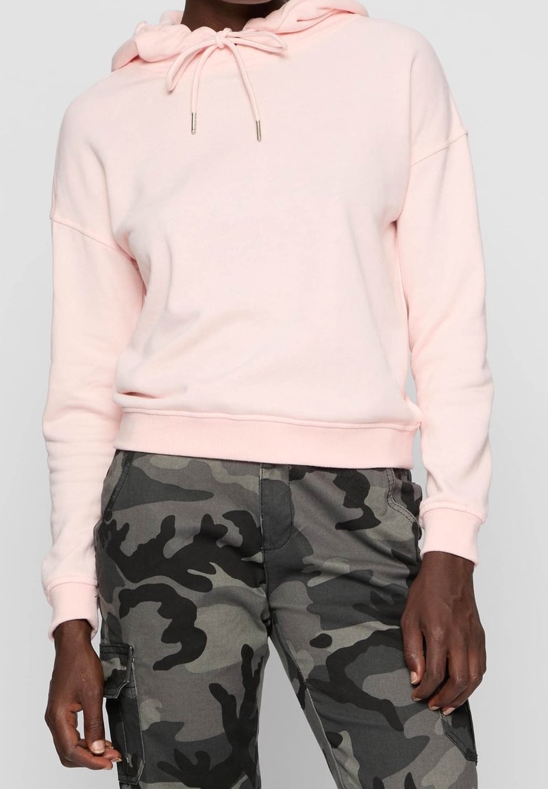 Personne portant un sweat à capuche rose clair et un pantalon cargo camouflage gris, debout les mains relâchées le long du corps.