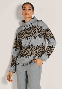 Frau in einem grauen Pullover mit braunen und schwarzen Leopardenmusterstreifen, hochgeschlossen, und grauen maßgeschneiderten Hosen, stehend mit einer Hand in der Tasche.