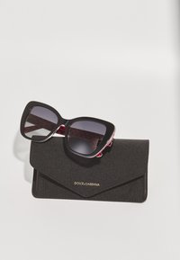 Black oversized sunglasses with a glossy finish, featuring dark lenses and a red heart pattern inside. Accompanied by a black case with a gold logo.