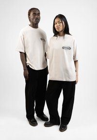 Two individuals stand side by side, wearing oversized beige t-shirts with black logo accents and black corduroy pants. They wear black shoes.