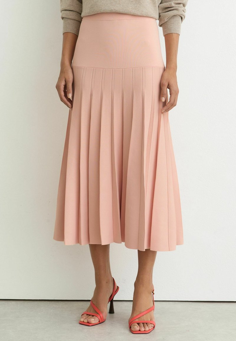 Femme portant une jupe midi plissée taille haute en rose doux et des sandales à talons hauts à lanières corail, debout contre un mur blanc.