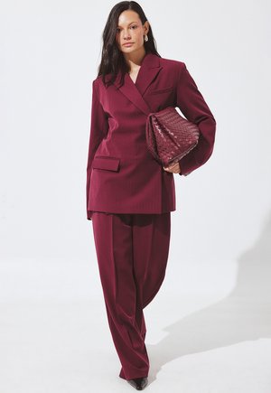 Completo sartoriale color bordeaux con blazer doppiopetto strutturato e pantaloni dal taglio morbido, abbinato a una borsa intrecciata dalla texture evidente e décolleté a punta.
