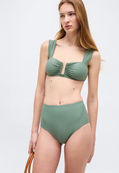 Γυναίκα με πράσινο μαγιό ψηλόμεσο με φαρδιές τιράντες στον ώμο, κρατώντας τη λαβή μιας καφέ τσάντας, μπροστά σε απλό ανοιχτό φόντο.