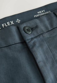 Nahaufnahme des taillierten dunkelschwarzen Hosenbunds mit einem schwarzen Knopf und sichtbarem Innenlabel mit der Aufschrift "NEXT PERFORMANCE FLEX".