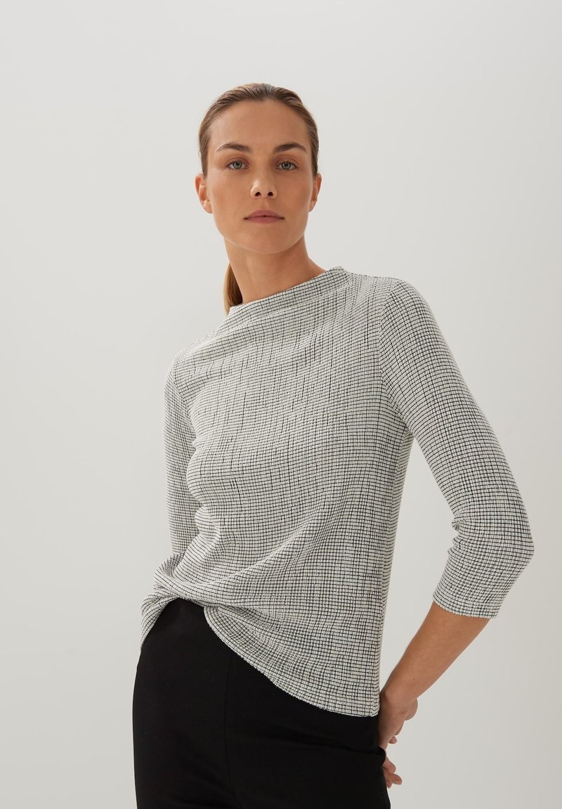 Schwarz-weißer gemusterter Oberteil mit Dreiviertelärmeln, rundem Ausschnitt und strukturiertem Stoff, getragen mit schwarzen Hosen.