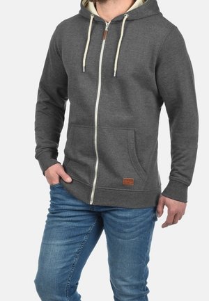 Homme portant un sweat à capuche zippé gris foncé avec des cordons crème et un jean bleu, posant avec une main dans la poche sur fond blanc.