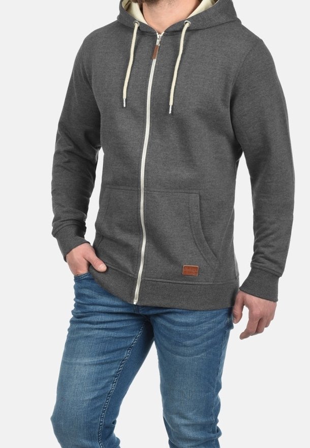 Homme portant un sweat à capuche zippé gris foncé avec des cordons crème et un jean bleu, posant avec une main dans la poche sur fond blanc.