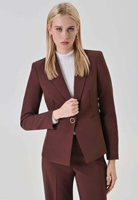 Blazer marrone su misura con chiusura a un bottone e revers a lancia. Indossato sopra una maglia bianca strutturata, presenta una vestibilità aderente e un tessuto liscio.