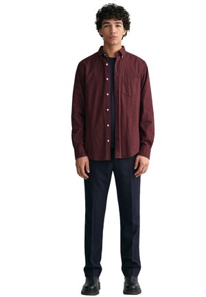 Camisa burdeos de botones con un bolsillo en el pecho, combinada con pantalones azul marino y zapatos negros. Tejido suave, mangas largas y forma ajustada.