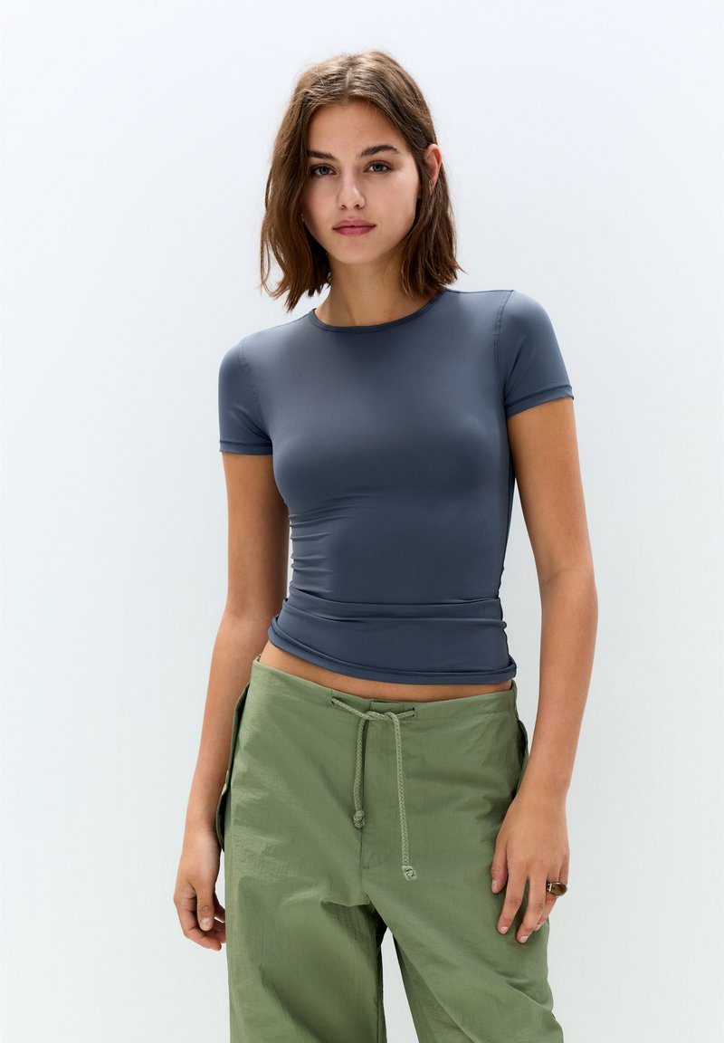 Prilega majica s kratkimi rokavi v sivi modri barvi, izdelana iz gladkega, raztegljivega materiala. V kombinaciji z ohlapnimi, olivno zelenimi hlačami s vrvico.