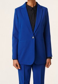 Osoba oblečená v královsky modrém obleku s odpovídajícím sako a kalhotami přes černou košili s knoflíky, stojící na jednobarevném pozadí.