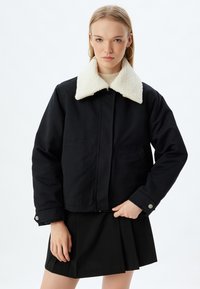 Schwarze Jacke mit einem weißen Kunstpelzkragen, vorderen Taschen und einem geknöpften Bund. Getragen über einem hellen Oberteil und einem schwarzen Rock.