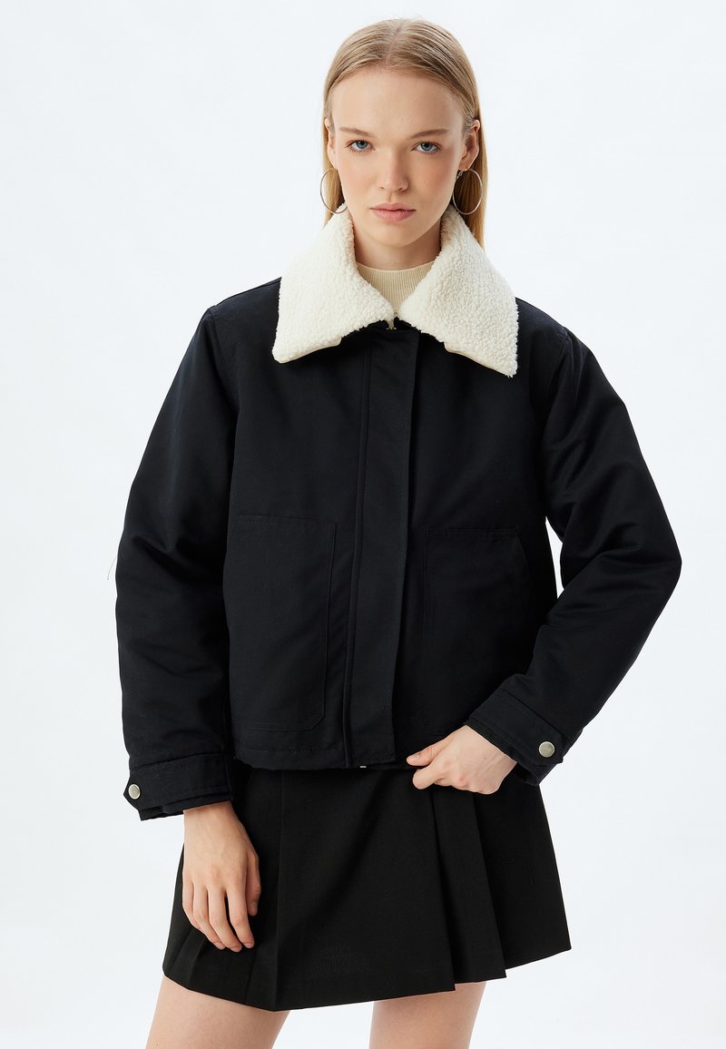 Schwarze Jacke mit einem weißen Kunstpelzkragen, vorderen Taschen und einem geknöpften Bund. Getragen über einem hellen Oberteil und einem schwarzen Rock.