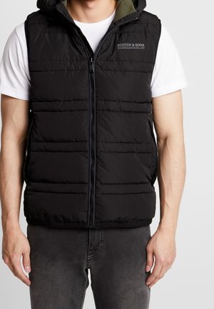 Mann trägt eine schwarze, gesteppte Steppweste mit Reißverschluss und Kapuze über einem weißen kurzärmligen Hemd und dunklen Jeans, steht vor einem einfarbigen Hintergrund.