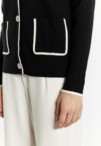Černý cardigan s bílým lemováním, s dvěma předními kapsami a zapínáním na knoflíky. Vyroben z měkkého materiálu, má dlouhé rukávy s akcentovými detaily.