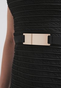 Robe noire texturée à rayures horizontales, avec une large ceinture noire ornée d'une boucle rectangulaire dorée brillante à la taille.