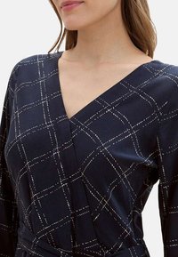 Robe portefeuille marine avec un motif à carreaux blancs, décolleté en V et manches longues. Fabriquée en tissu léger avec une texture lisse.