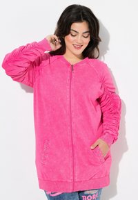 Sudadera rosa con cremallera y mangas texturizadas, puños acanalados y detalles de tachuelas en los bolsillos. Tejido suave y ligero.